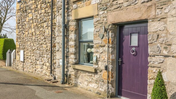 A stone building with a purple door, a window with decorations, and a small window above the door. - Bank End Cottage (Carnforth)