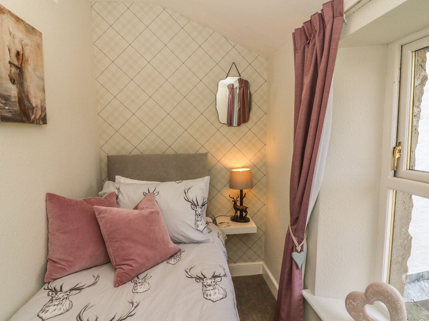 A bedroom with a bed, pillows, a nightstand, a lamp, a mirror, and a window with curtains.