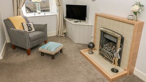 A cozy living room with a striped armchair, a small ottoman, a fireplace, and a television on a white cabinet. - Bank End Cottage (Carnforth)