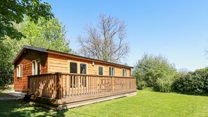 A wooden cabin with a deck, surrounded by a well-maintained lawn and trees. - Lodge One (Truro)