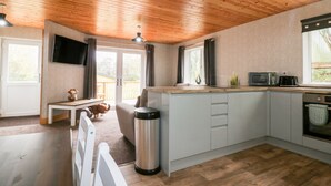 A compact kitchen with a wooden ceiling, a TV mounted above the counter, and a window overlooking a deck. - Lodge One (Truro)