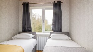 A bedroom with two beds, a window with curtains, and a radiator. - Lodge One (Truro)