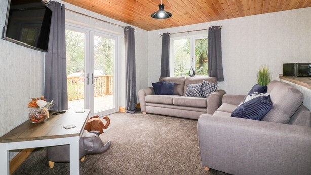 A cozy living room with a wooden ceiling, a grey sofa set, a dining table with a remote control, and a TV mounted on the wall. - Lodge One (Truro)