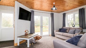 A modern living room with a wooden ceiling, a flat-screen TV mounted on the wall, a sofa, a coffee table, and a large window with curtains. - Lodge One (Truro)