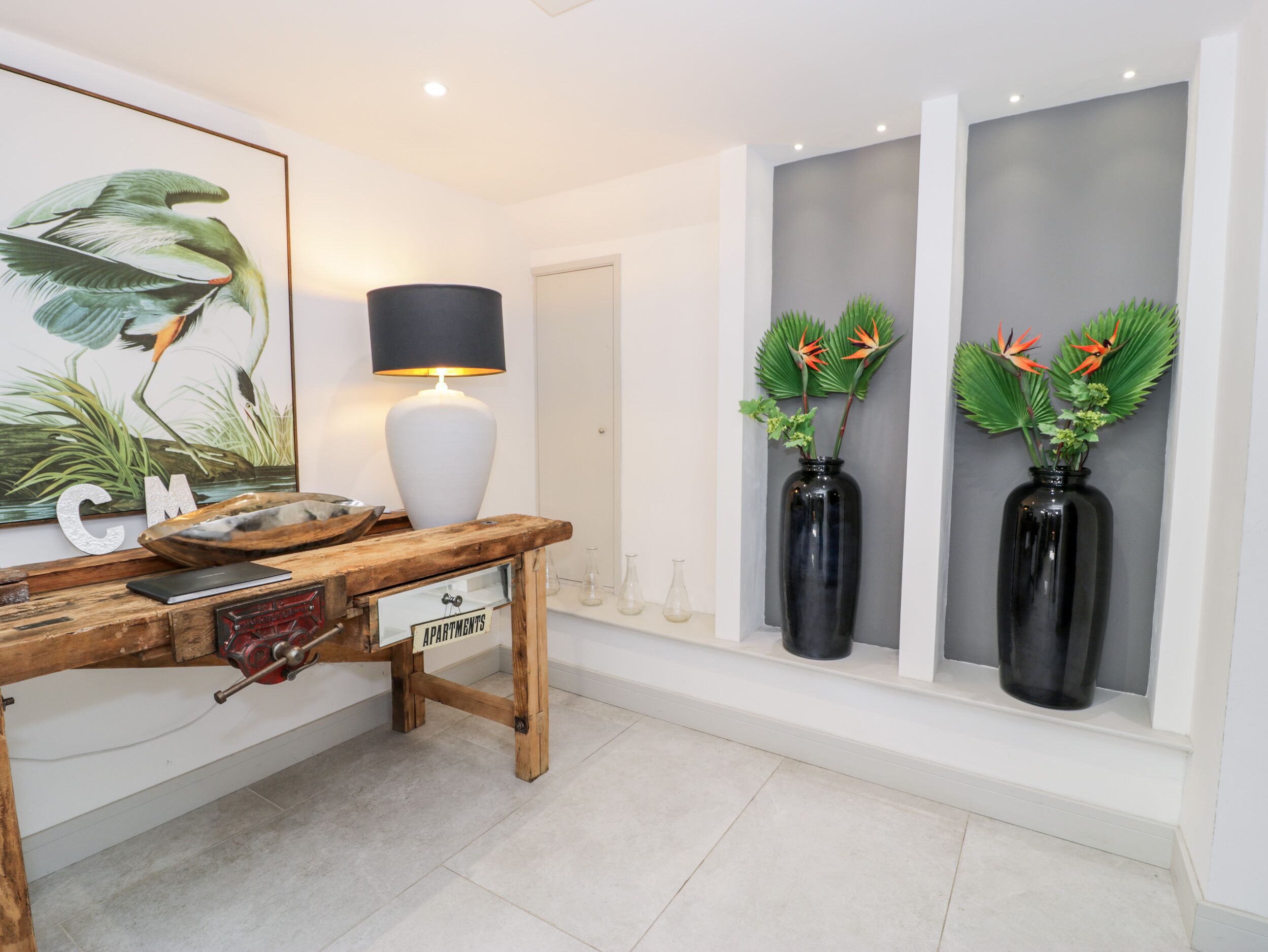 A modern interior with a wooden desk, a framed artwork, a lamp, and two black vases with decorative plants.