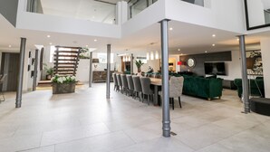 A spacious, modern interior with a high ceiling, a dining area with multiple chairs, a staircase, and a living area with a green sofa and a flat-screen TV.