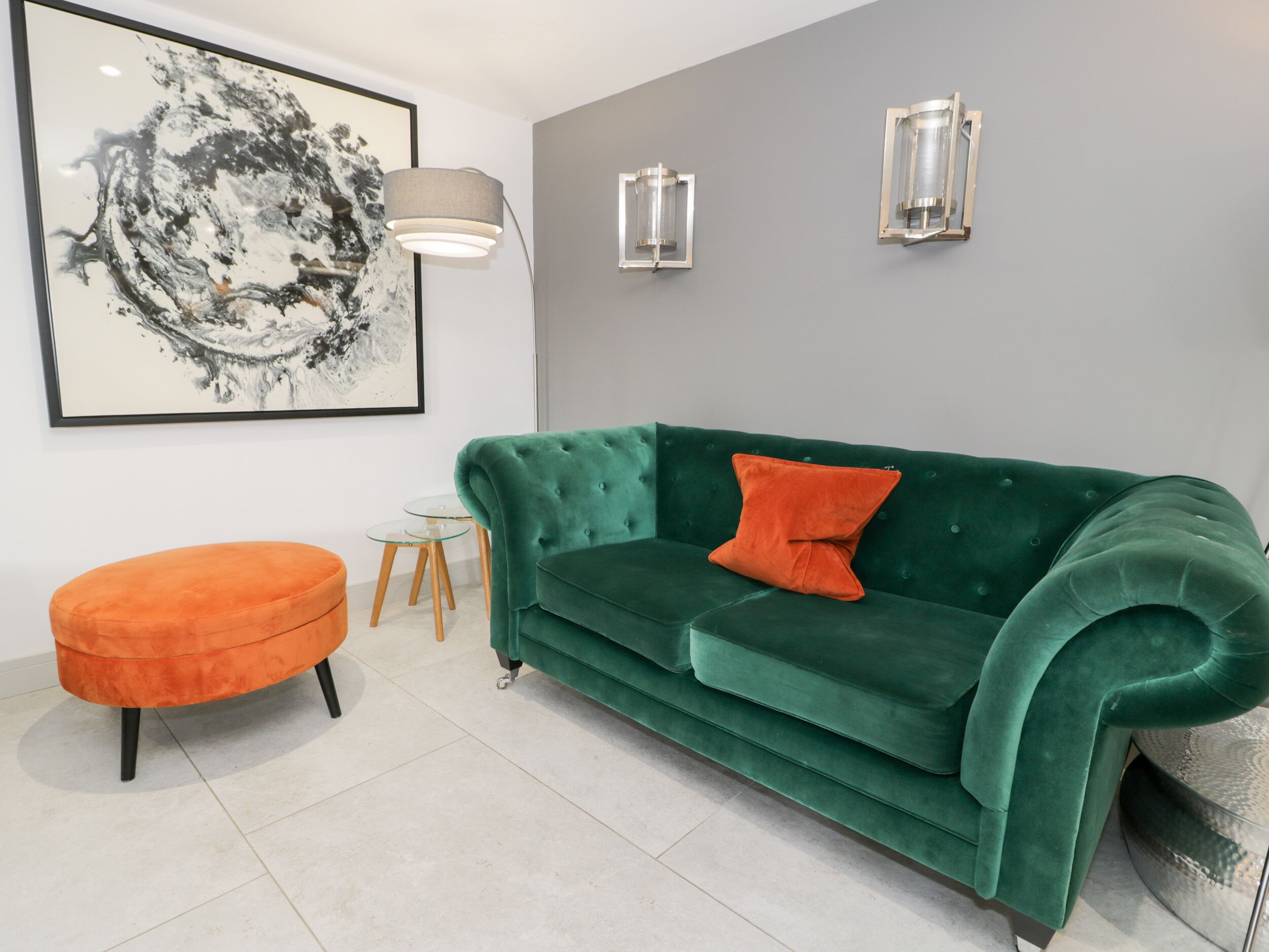 Un salon avec un canapé en velours vert, un pouf orange, une petite table ronde et un tableau abstrait encadré accroché au mur.