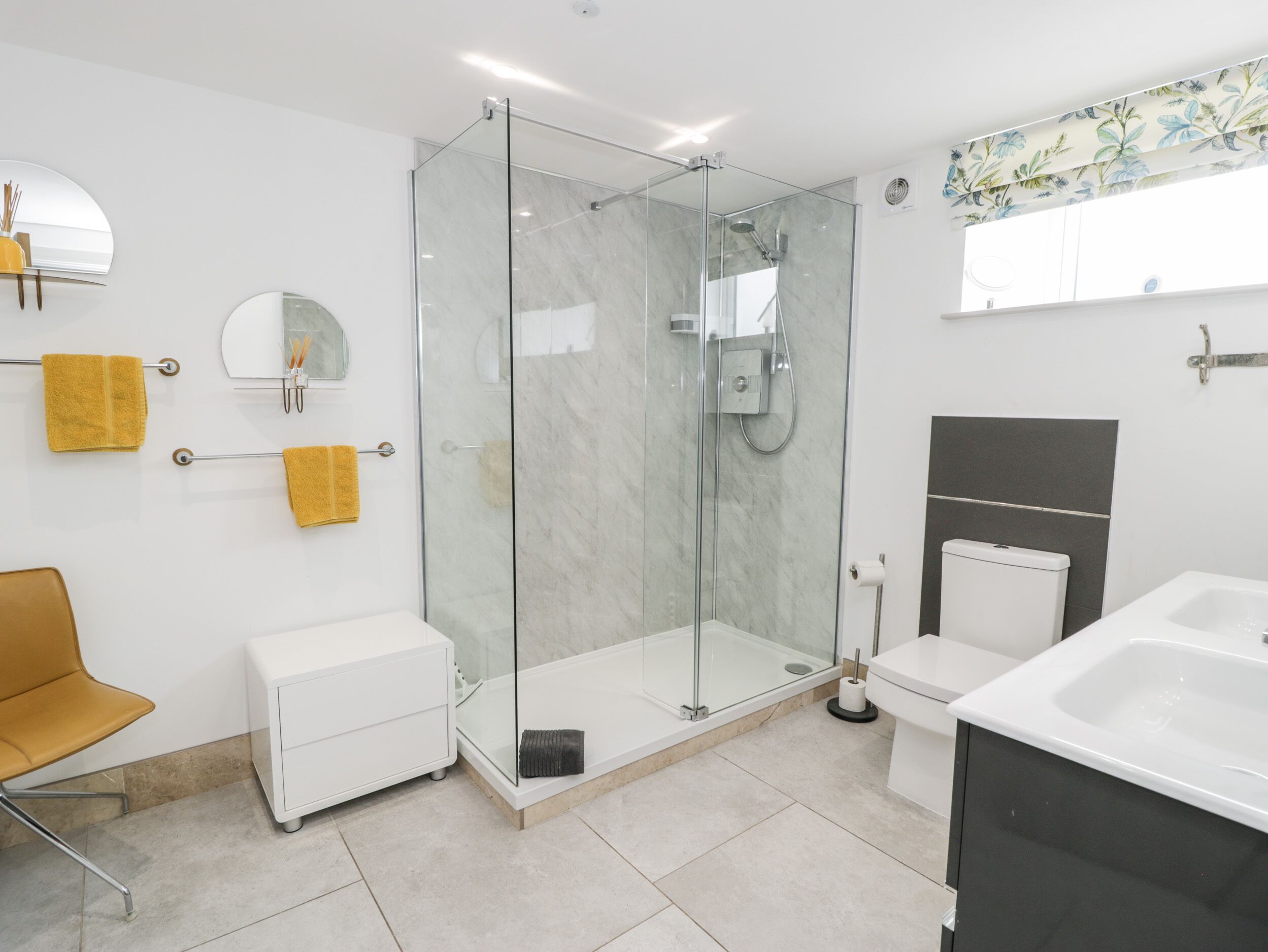 Un bany modern amb una dutxa envidriada, un lavabo blanc i un vàter. Hi ha una tovallola groga sobre un suport i un mirall a la paret.