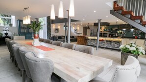A spacious dining area with a long table and multiple chairs, a kitchen in the background, and a staircase leading to an upper level.
