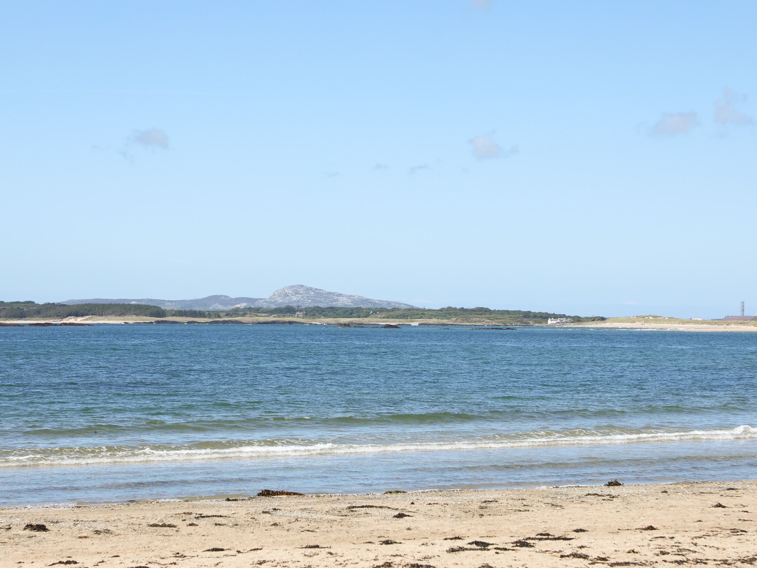 A beach with clear blue water, sandy shore, and distant hills.