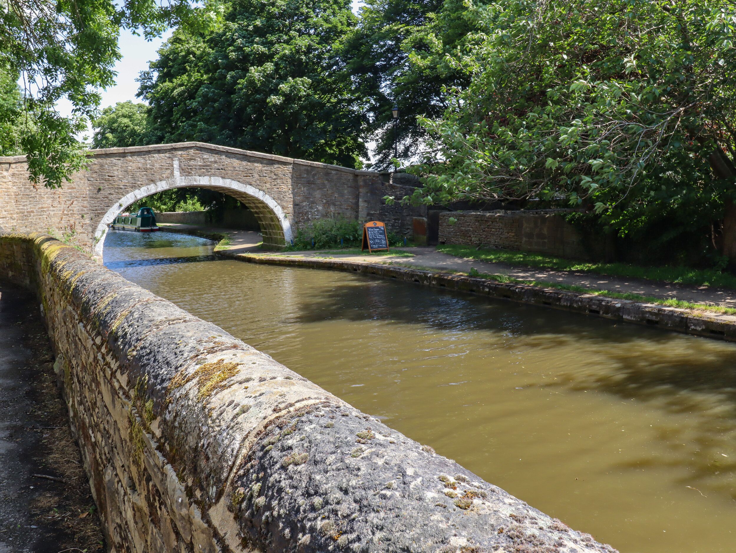 A stone bridge over a narrow canal with a signboard and lush greenery in the background.