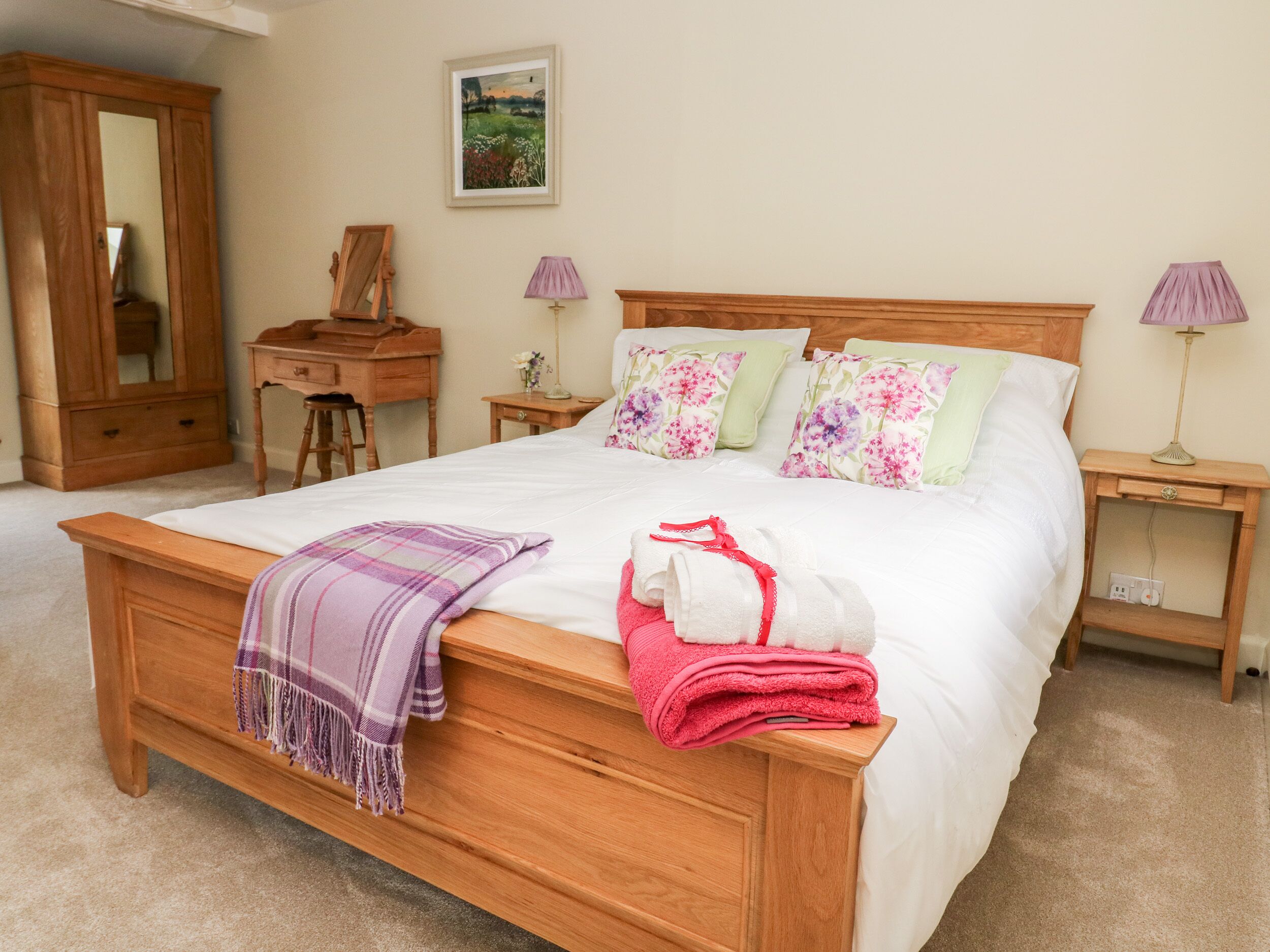 A neatly made bed with a plaid throw, a wooden nightstand with a lamp, a wooden dresser, and a desk with a mirror.