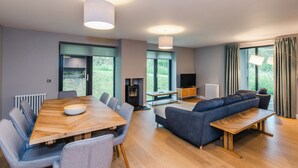 A modern living room with a wooden dining table, grey upholstered chairs, a blue sofa, and a television. - Mountain Hare Apartment (Kingussie)