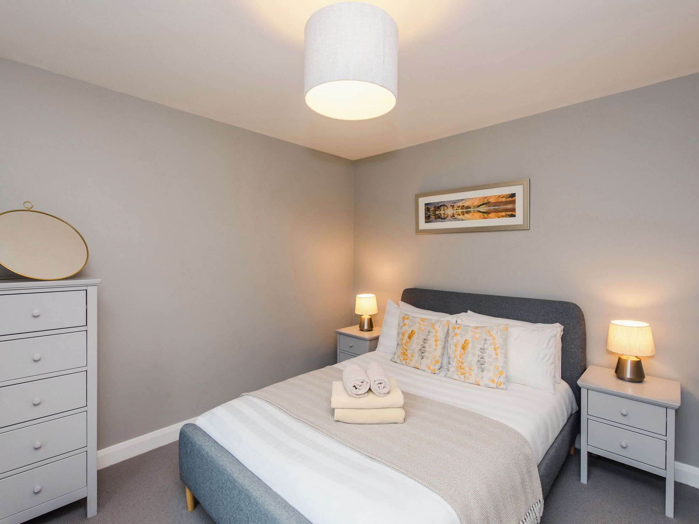Un dormitorio ordenado con una cama, mesitas de noche, una lámpara, un espejo y un cuadro en la pared.
