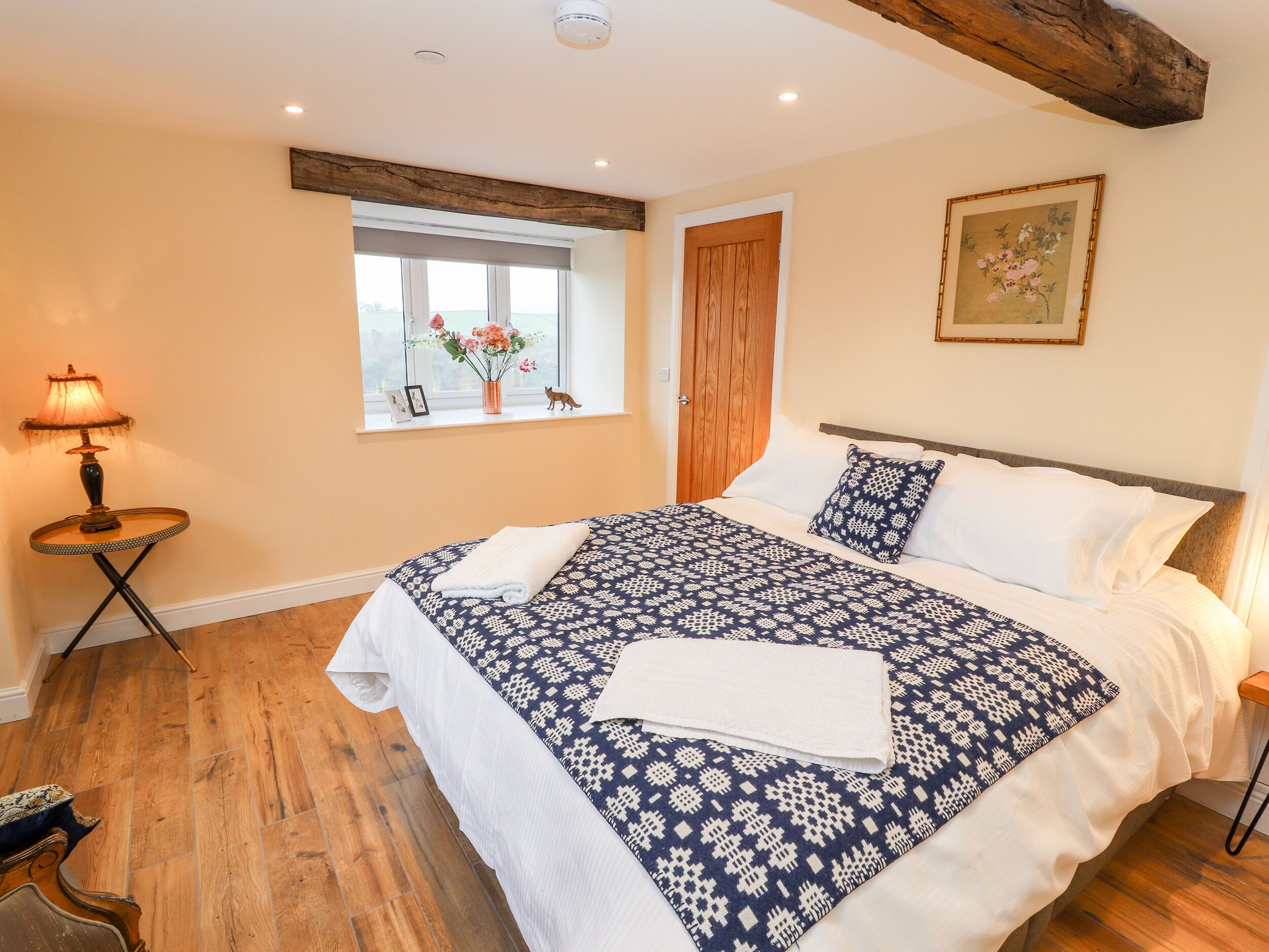 A bedroom with a large bed, a window with flowers, a wooden door, and a small table with a lamp.