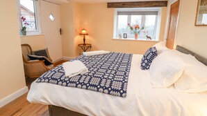 Un dormitorio con una cama grande, una mesita de noche con lámpara, una ventana con flores y una silla.