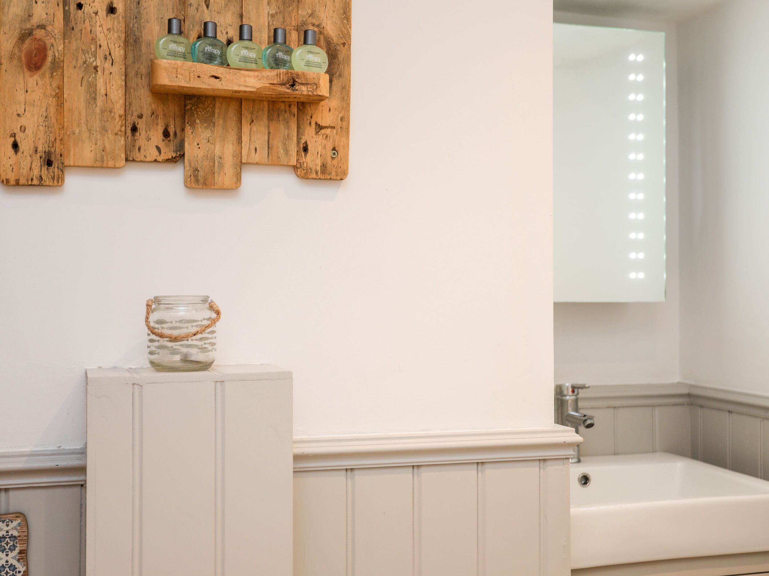 A bathroom with a wooden shelf holding green bottles, a glass jar with a rope handle, and a large mirror with lights.