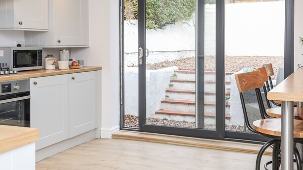 A modern kitchen with white cabinets, a microwave, and a coffee maker. There is a sliding glass door leading to an outdoor area with steps and a garden.