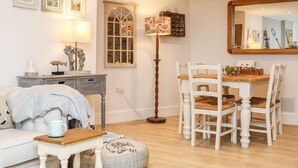 A cozy living room with a white sofa, a wooden coffee table, and a dining table with white chairs. There is a floor lamp, a mirror, and framed artwork on the wall.
