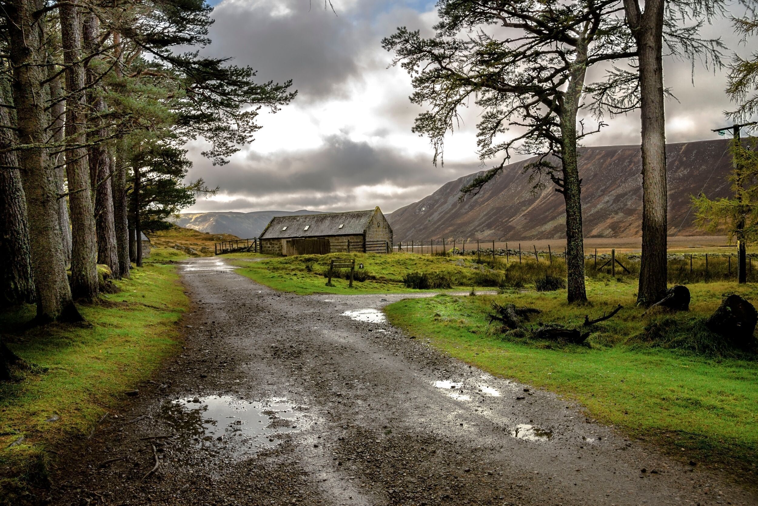 A rural road leading to a stone barn with a fenced pasture and mountains in the background.
