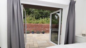 Ein Schlafzimmer mit einem Bett, einem weißen Nachttisch und einem Beistelltisch. Der Raum hat eine Glasschiebetür, die auf eine Terrasse mit einer Backsteinmauer, Pflanzgefäßen und einer Sitzecke führt.