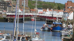 Ein Hafen mit vertäuten Booten, eine Brücke im Hintergrund und Gebäude entlang der Uferlinie.