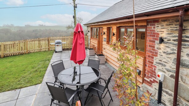 A patio with a table and chairs, a red umbrella, and a view of a stone building with a wooden deck. - Cut Moch, Plas Moelfre Hall Barns (Oswestry)
