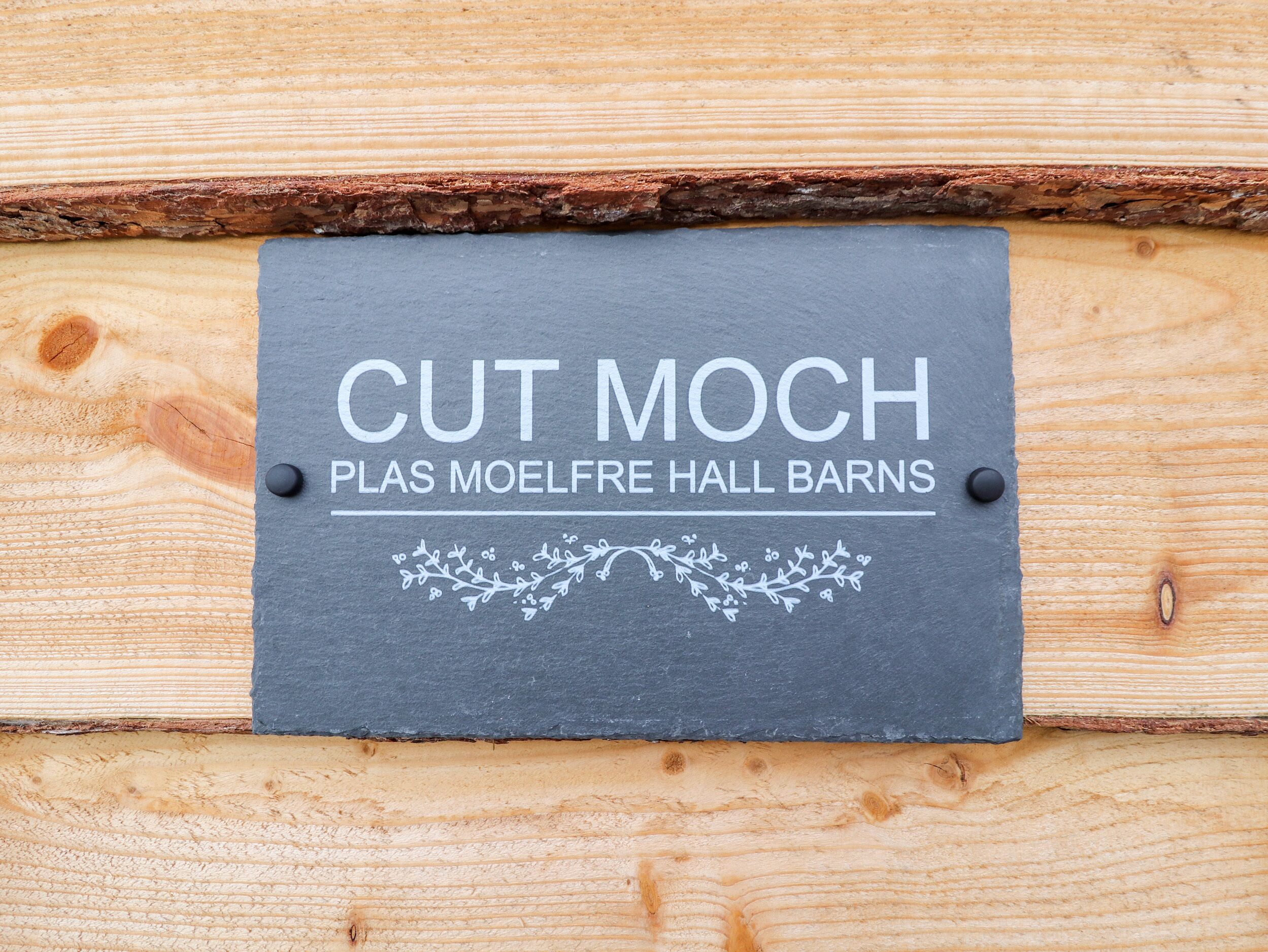 Un letrero de pizarra con la inscripción 'CUT MOCH' y 'PLAS MOELFRE HALL BARNS', sujeto a una pared de madera.