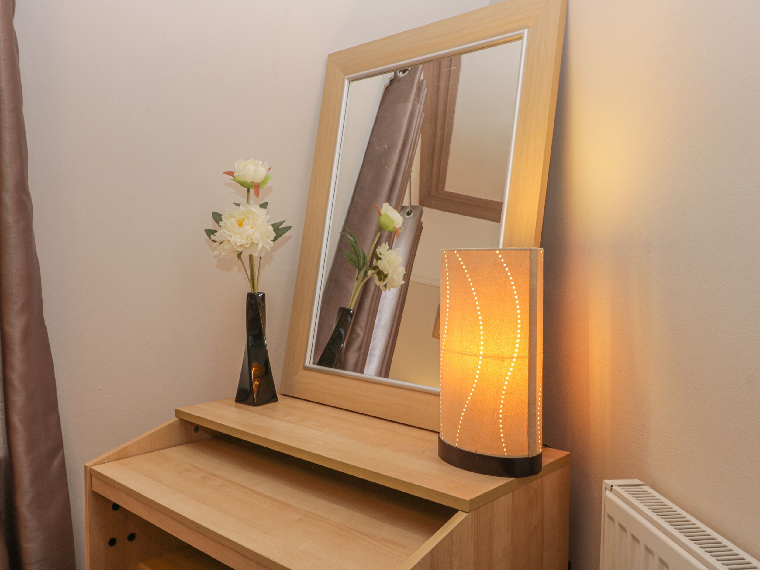 A wooden bedside table with a mirror, a vase with flowers, and a wall-mounted light fixture.