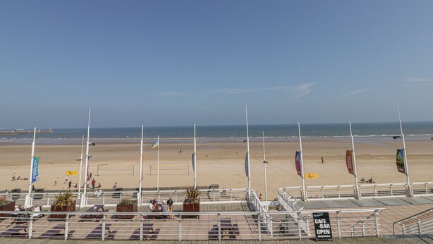 A beach view with a boardwalk, flags, and a sign indicating a cafe bar open downstairs. - Cosy Coast Cottage (Bridlington)
