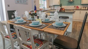 A dining area with a wooden table set for four, surrounded by white chairs with colorful seat cushions. A kitchen with white cabinets and a blue accent wall is visible in the background. - Cosy Coast Cottage (Bridlington)