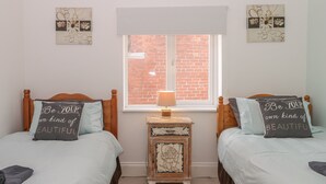 A bedroom with two beds, each with a pillow that says "Be your own kind of beautiful." There's a small wooden nightstand with a lamp and a picture frame between the beds. A window with a view of a brick wall is above the nightstand. - Cosy Coast Cottage (Bridlington)