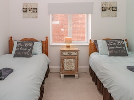 Un dormitorio con dos camas, cada una con una almohada que dice 'Be your own kind of beautiful'. Hay una pequeña mesita de madera con una lámpara y un marco de fotos entre las camas. Una ventana con vista a un muro de ladrillos está sobre la mesita.