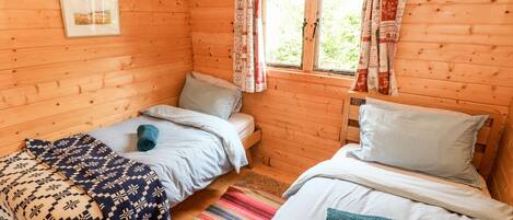 A wooden cabin room with two beds, a window with curtains, a framed picture on the wall, and a colorful striped rug.