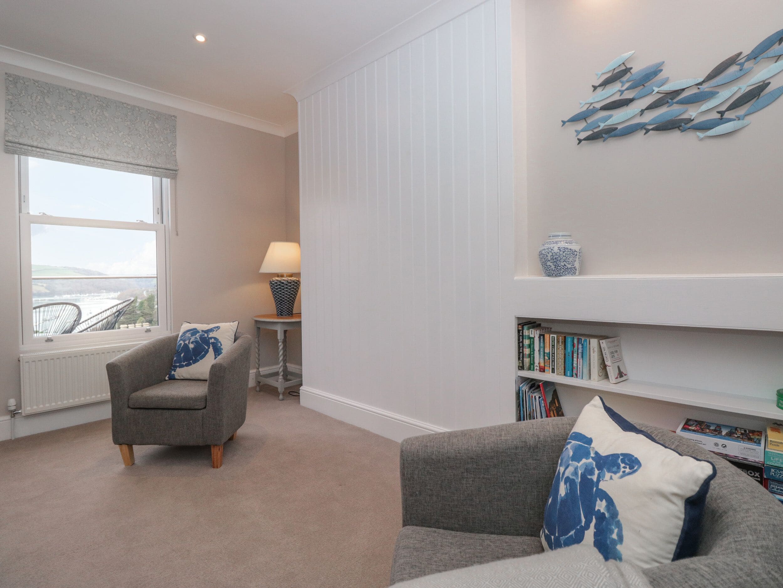 A living room with a grey sofa, a smaller armchair, a bookshelf, a window with a view, and a decorative fish wall art piece.