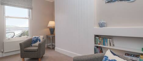 A living room with a grey sofa, a smaller armchair, a bookshelf, a window with a view, and a decorative fish wall art piece.
