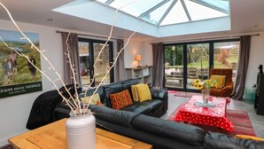 A modern living room with a black leather sofa, a wooden coffee table, and a glass-top dining table with a red tablecloth. There is a large window with curtains, a skylight, and a poster on the wall. - Carr House Farm (Glossop)
