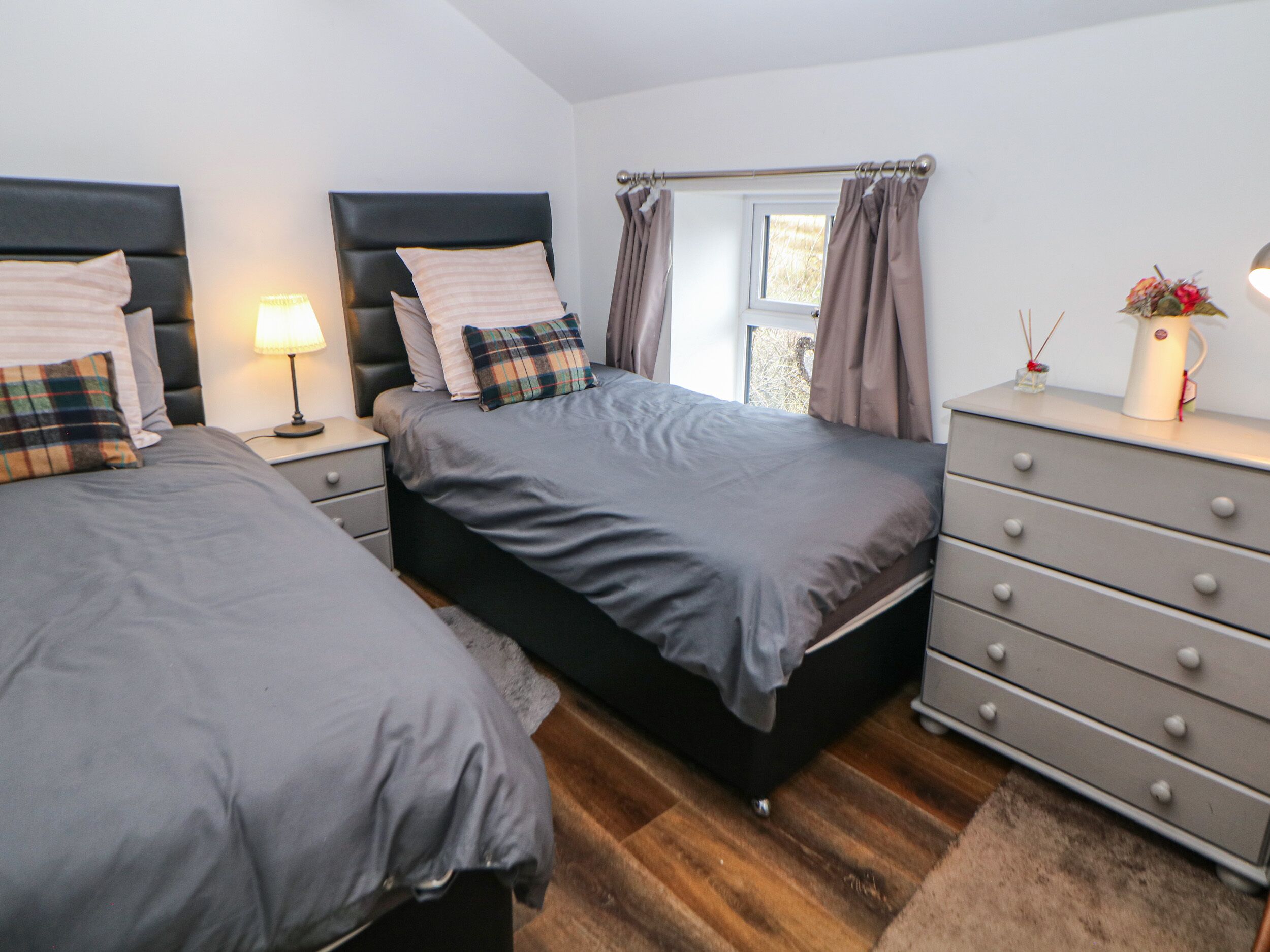 A bedroom with two beds, a bedside table with a lamp, a dresser, and a window with curtains.