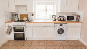 Una cocina compacta con armarios blancos, encimera de madera, estufa, microondas, lavadora y ventana con persianas.