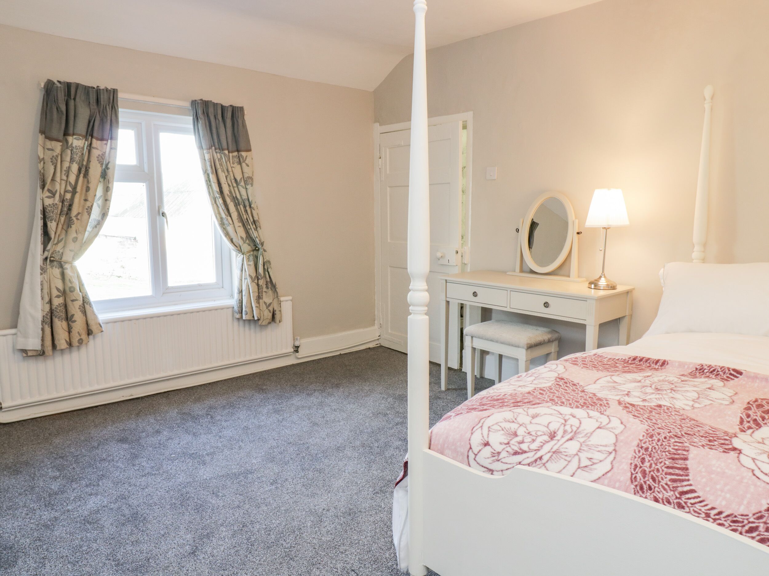 A bedroom with a four-poster bed, a bedside table with a lamp, a dresser, and a window with curtains.