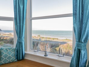A room with a large window overlooking a coastal road and ocean. - Llys Madoc, First Floor Flat (Abergele)