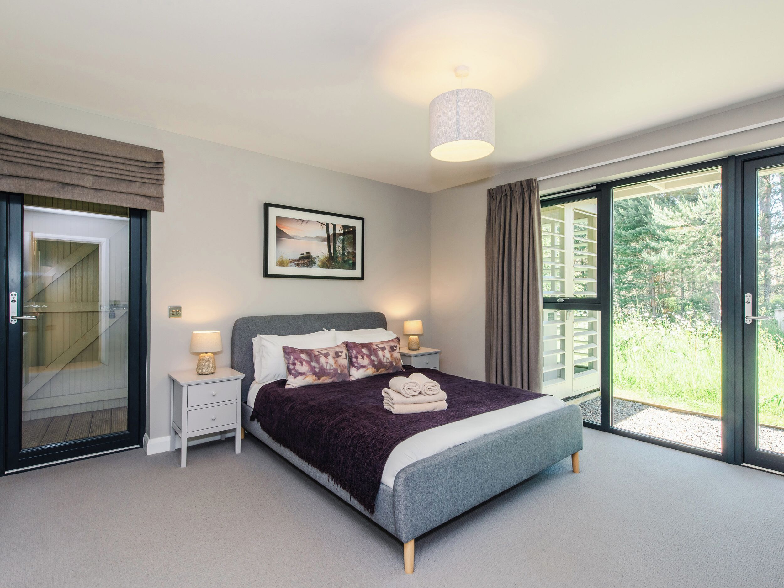 A modern bedroom with a large bed, two bedside tables with lamps, a framed picture on the wall, and a sliding glass door leading to a balcony.