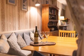 A wooden dining table with a bottle of wine and two glasses on a coaster, a wooden chair, and a bookshelf in the background. - Old Post Office (Amlwch)