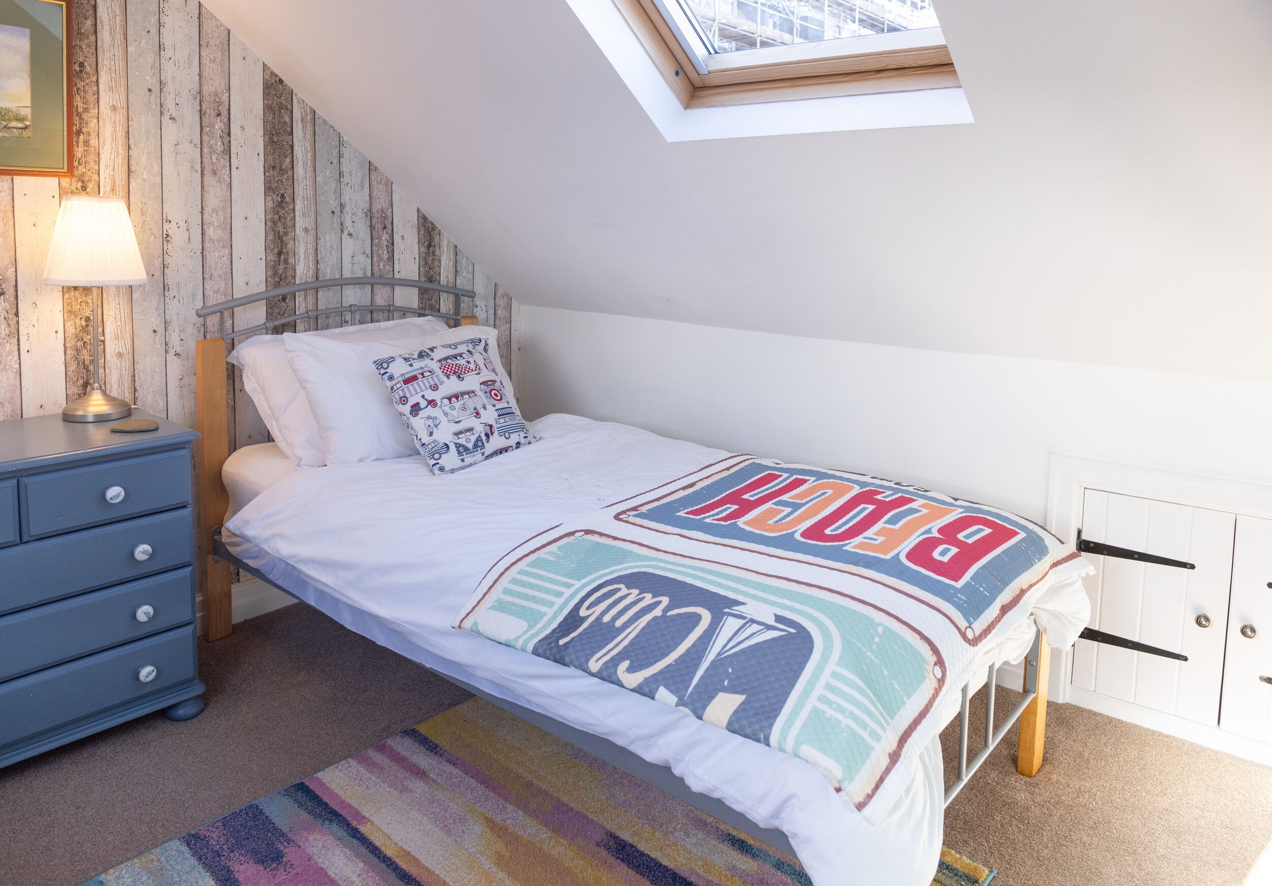 A bedroom with a sloped ceiling, a bed with a colorful bedspread, a nightstand with a lamp, a dresser, and a window with a view of the sky.