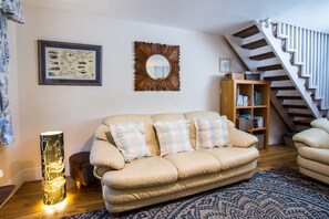 A living room with a beige sofa, a patterned rug, a wooden bookshelf, and a staircase. - Old Post Office (Amlwch)