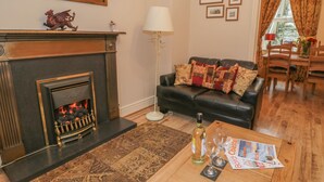 Uma sala de estar com lareira, um sofá de couro preto, uma mesa de centro de madeira com revistas e copos, e um espaço de jantar com cadeiras e mesa de madeira.