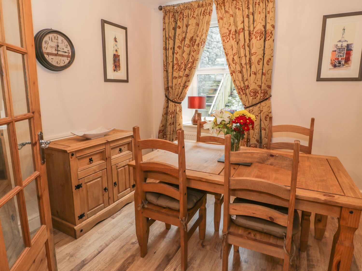 A wooden dining table with four chairs, a wooden cabinet with drawers, a wall clock, and a window with curtains.