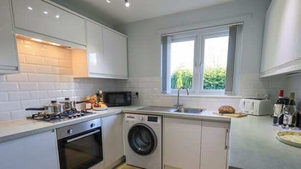 A compact kitchen with a washing machine, microwave, oven, and a window with a view of greenery. - Fairfell (Keswick)