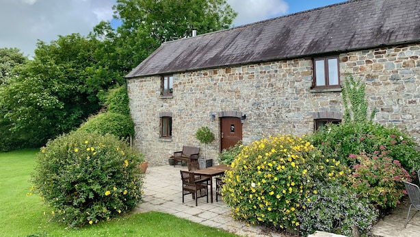A stone building with a tiled patio, wooden furniture, and flowering plants. - The Granary (Cardigan)