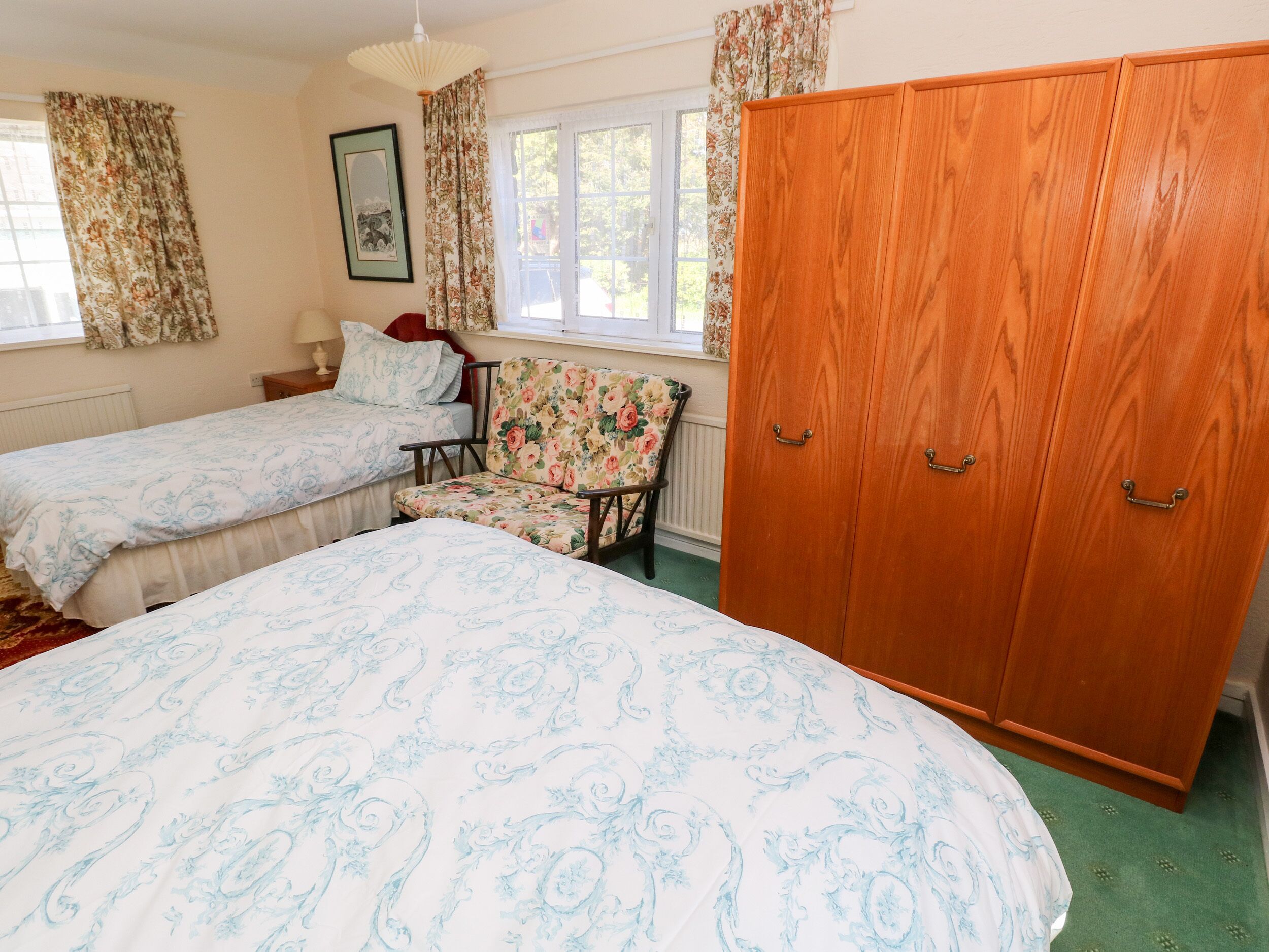 A bedroom with two beds, a wooden wardrobe, a floral chair, and a window with curtains.